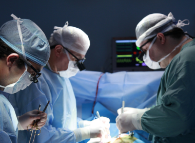 Surgeons in an operating room performing a live medical procedure with monitoring equipment in the background.